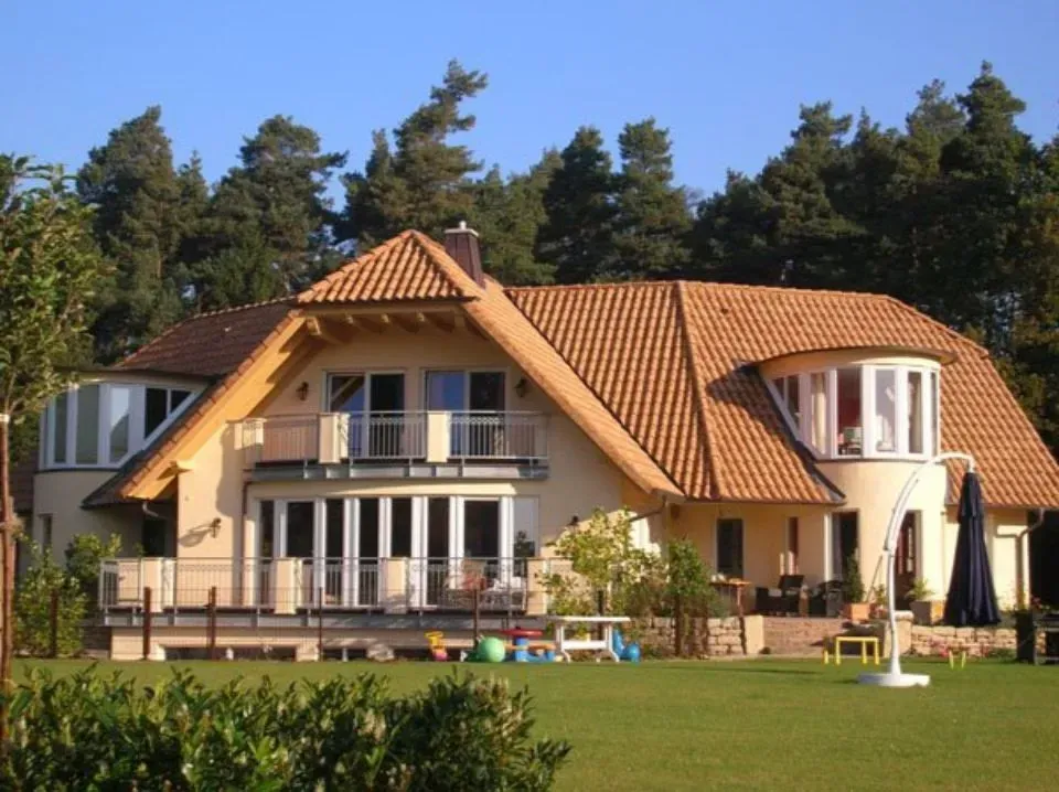 Haus mit rotem Ziegeldach und Terrasse, eingebettet in einen grasbewachsenen Garten mit dahinterliegenden Bäumen.