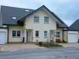 Vorstadthaus mit beiger Fassade, dunklem Dach, Garage und Vorgarten zur Straße hin