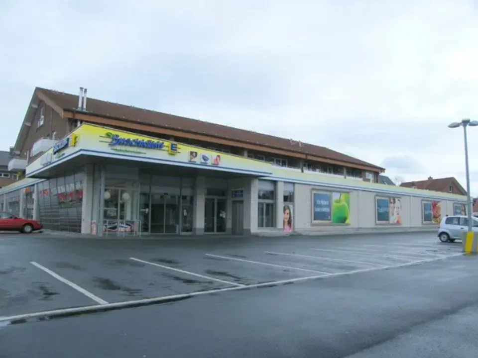 Ein kleiner Laden am Straßenrand mit gläsernem Eingang, gelbem Schild und Parkplatz an einem bewölkten Tag.