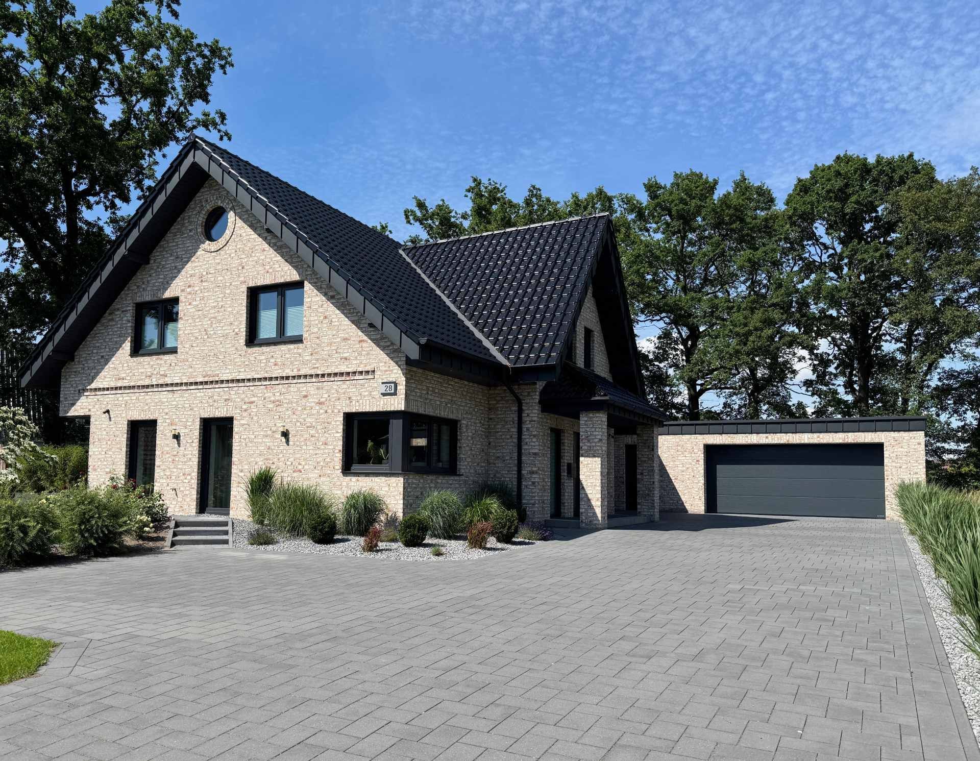 Steinhaus mit dunklem Dach und angebauter Garage, von einer gepflasterten Auffahrt an einem sonnigen Tag aus betrachtet.