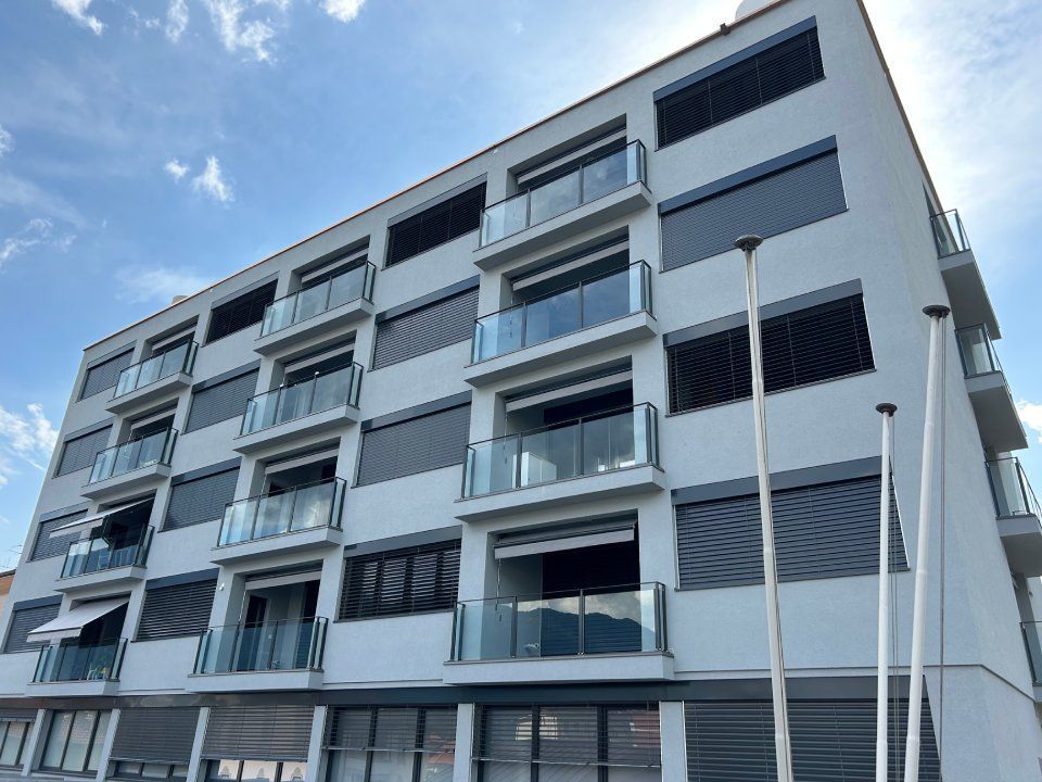 Moderno edificio a più piani con balconi, ringhiere in vetro e tende alle finestre, contro un cielo nuvoloso.