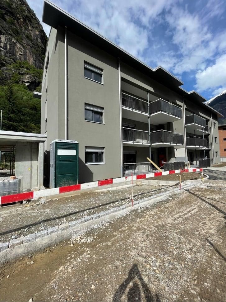 Nuovo condominio grigio in costruzione, con balconi, una montagna sullo sfondo e barriere edili di fronte.