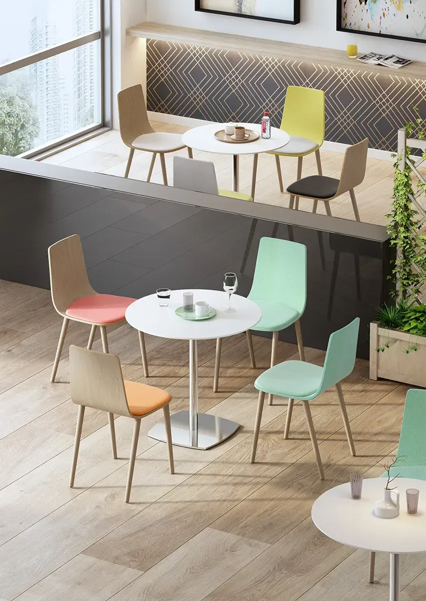 Ambiente de cafetería con mesas redondas blancas, sillas de colores y una ventana.