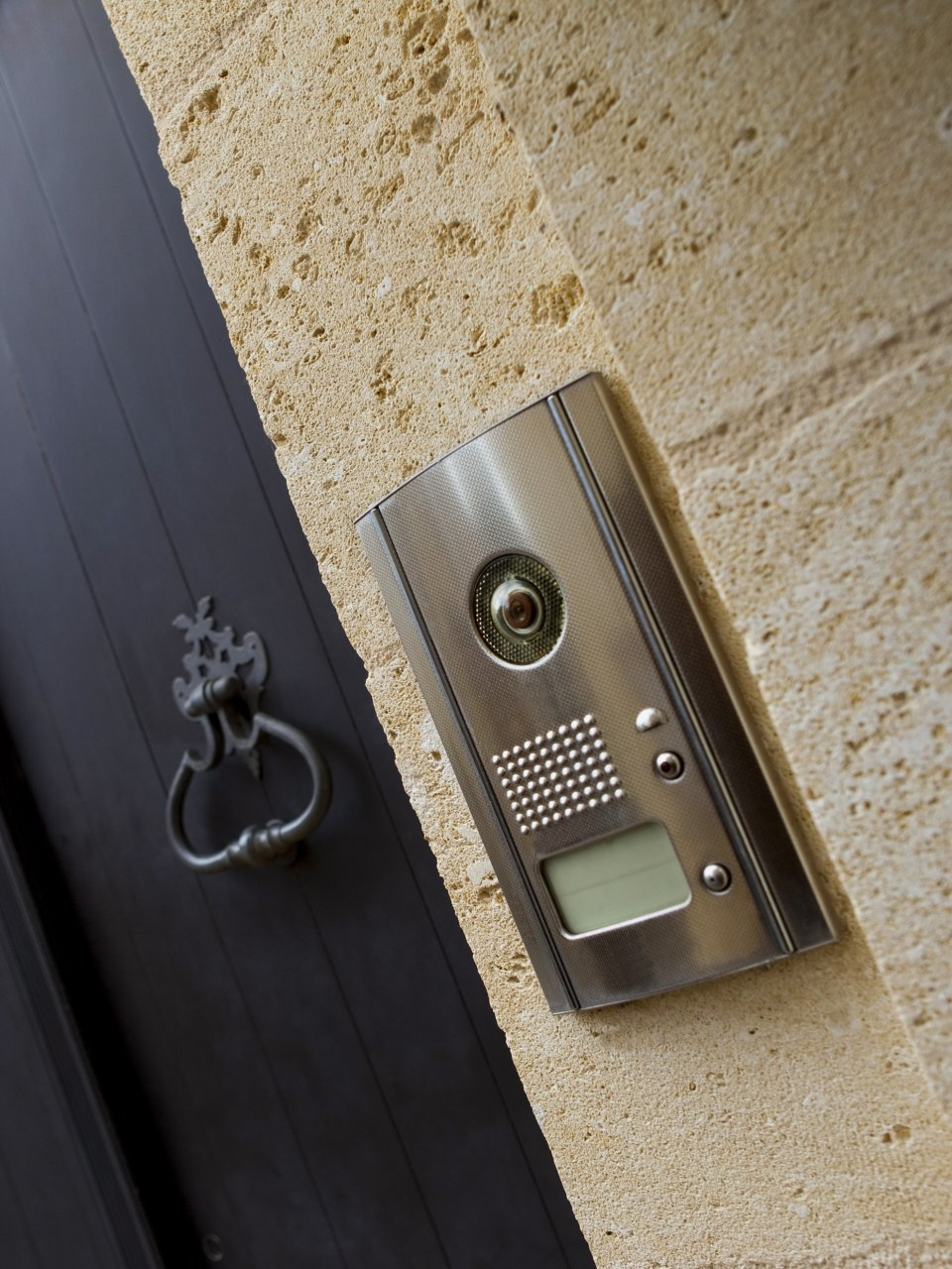 Un panneau d'interphone en acier inoxydable fixé sur un mur en pierre beige, à côté d'une porte noire munie d'un heurtoir décoratif.