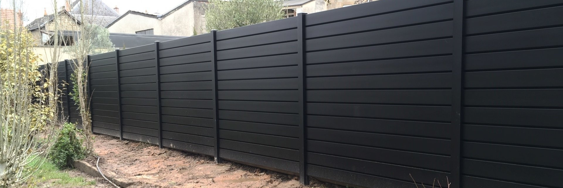 Une longue clôture sombre à lattes horizontales se dresse devant une maison. Végétation au premier plan.