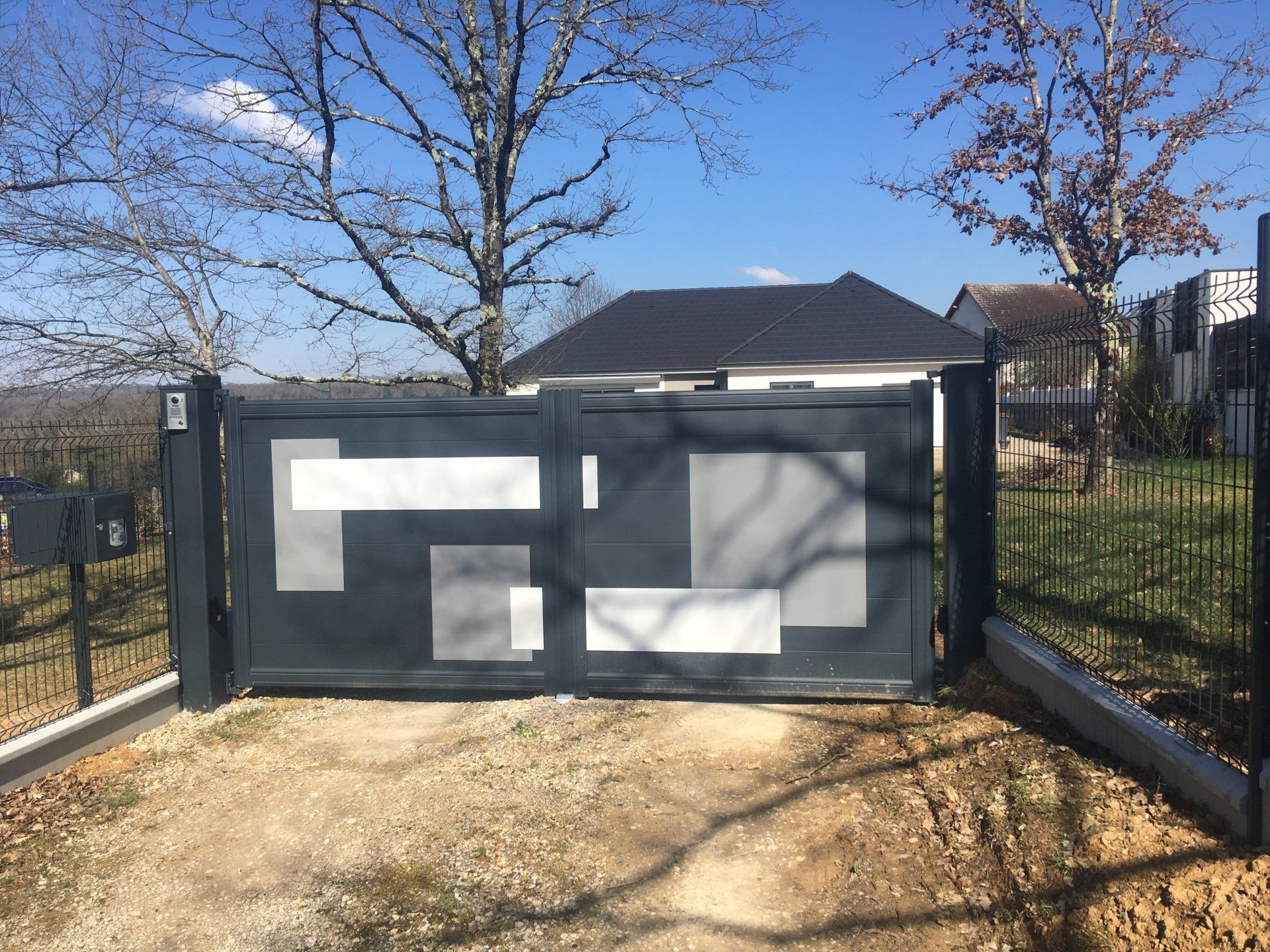 Portail moderne gris foncé à motif géométrique, devant une maison donnant sur une allée de gravier.
