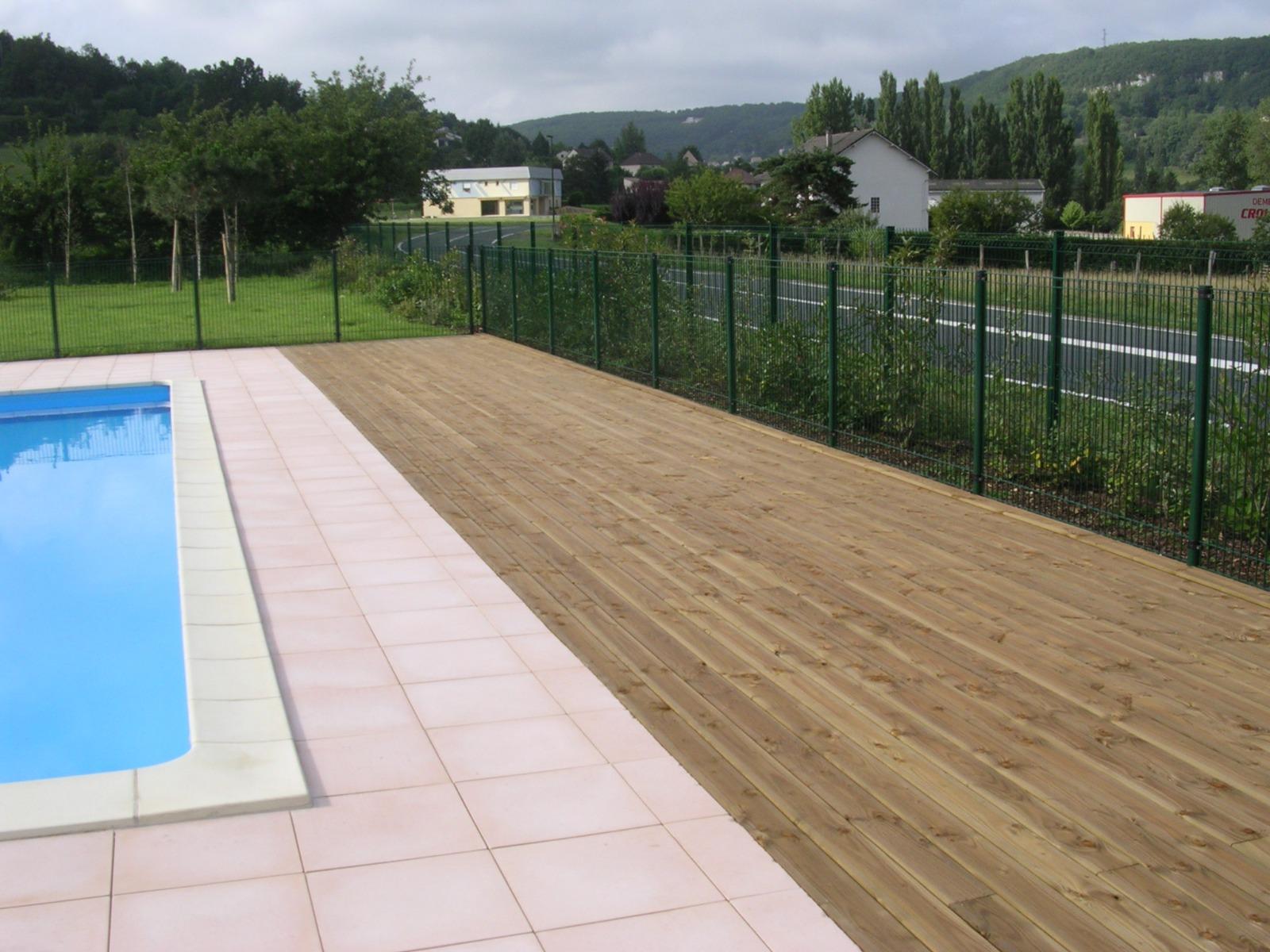 Bord de piscine avec terrasse carrelée, terrasse en bois, clôture verte et arrière-plan d'arbres et de maisons.