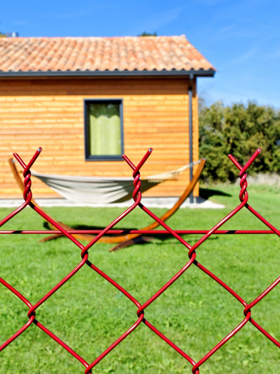 Clôture en grillage rouge au premier plan ; cabane en bois et hamac en arrière-plan sur l'herbe verte, journée ensoleillée.