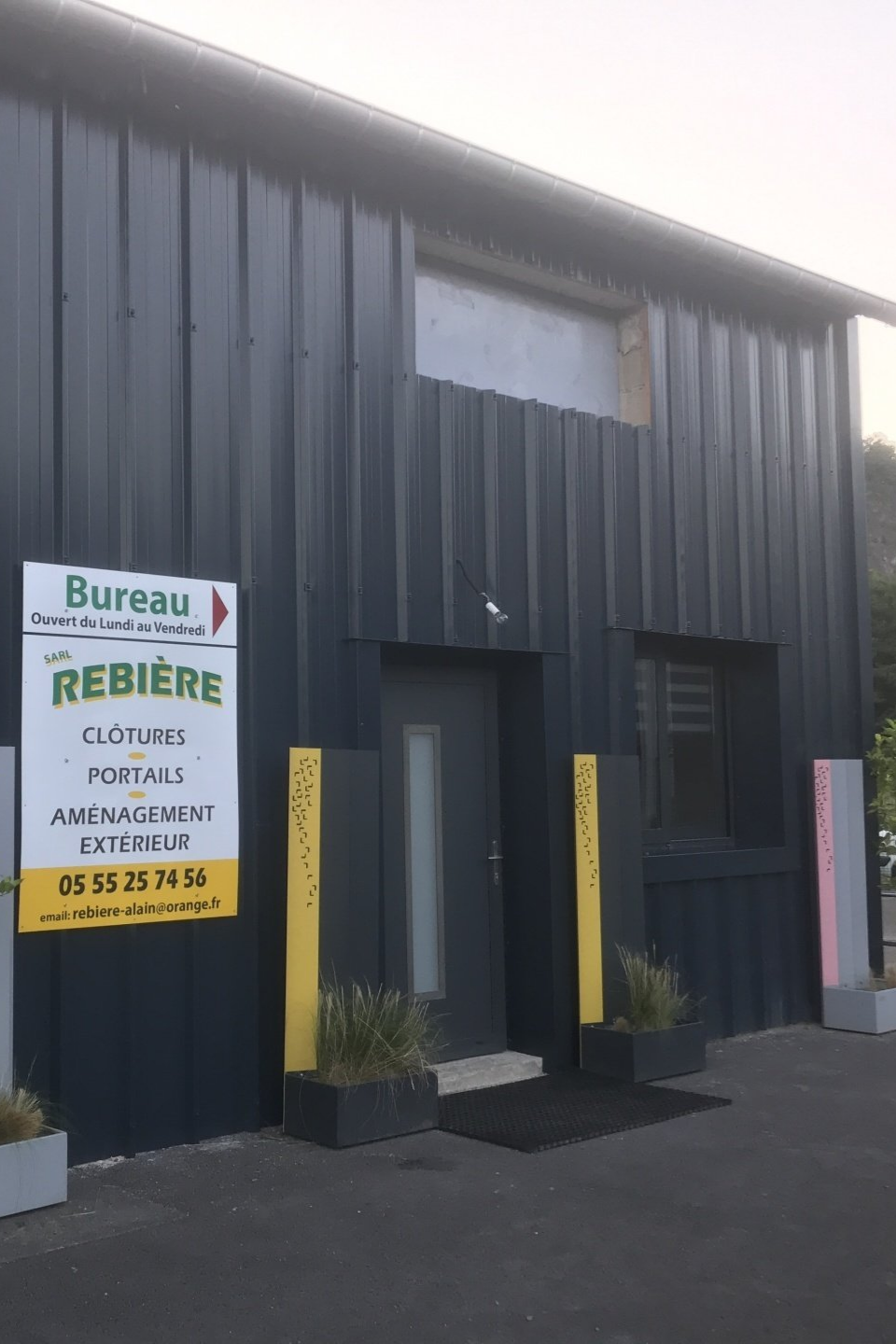 Bâtiment gris foncé avec entrée, enseigne publicitaire, touches de jaune et plantes en pot.