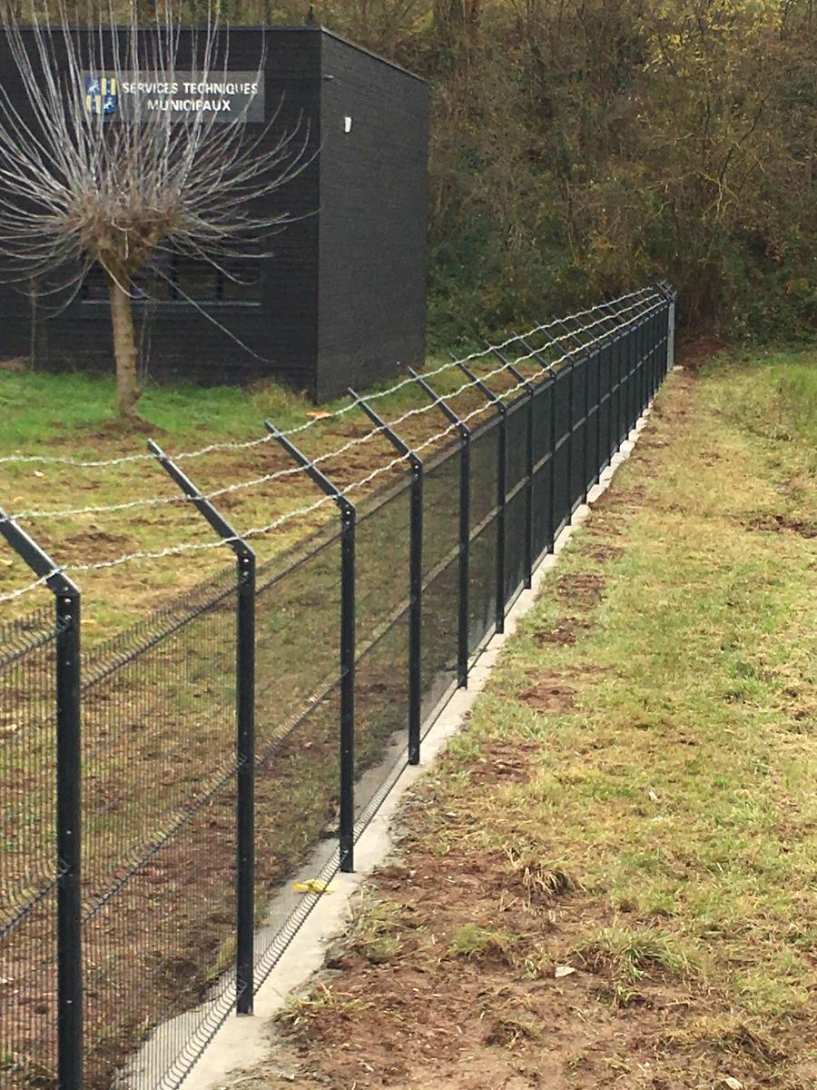 Clôture en fil de fer noir hérissée de pointes le long d'un champ herbeux, à côté d'un bâtiment noir.