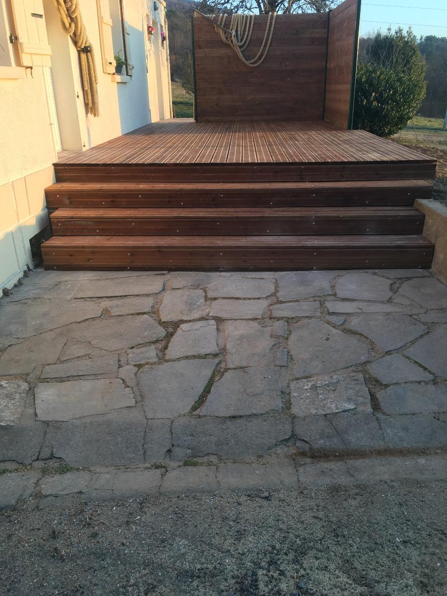 Des marches en bois mènent à une terrasse en bois, située sur un patio en pierre, à côté d'un bâtiment.