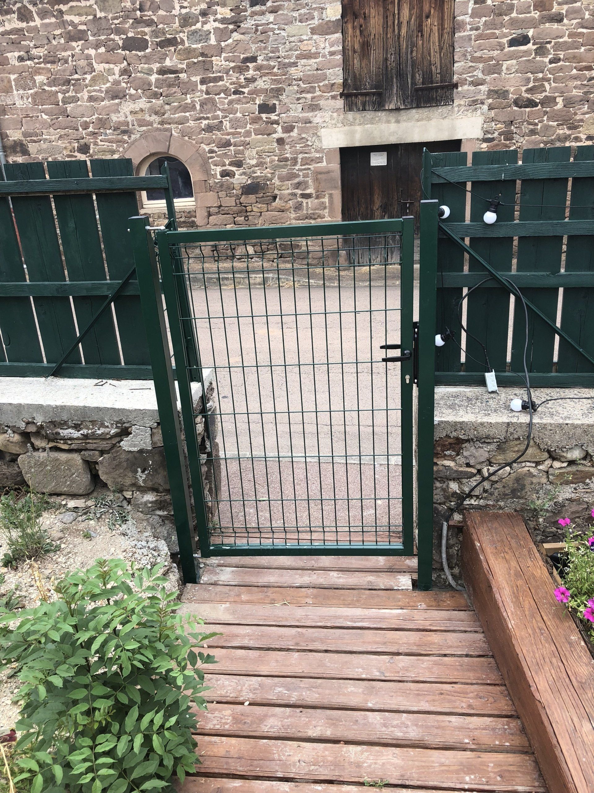 Portail en métal vert dans un mur de pierre, un chemin en bois y mène.