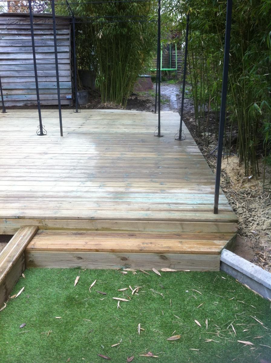 Terrasse en bois avec marches dans un jardin, bordée de gazon synthétique. Des supports en métal noir émergent de la terrasse.