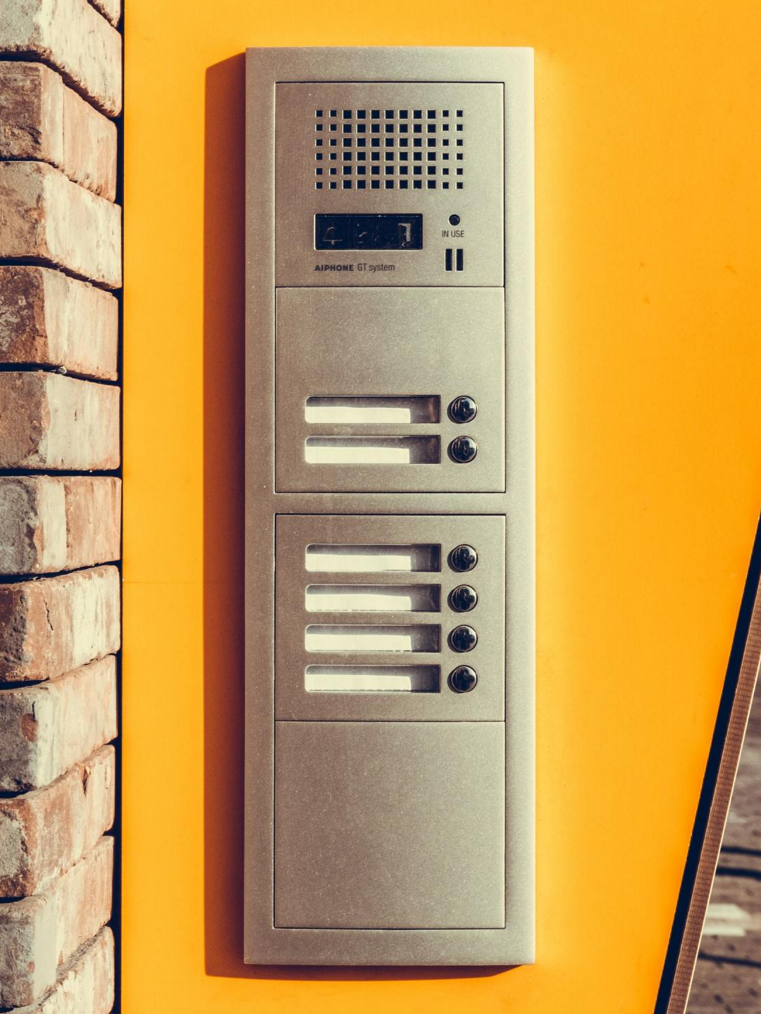 Panneau d'interphone argenté avec boutons et haut-parleur sur un mur orange à côté d'une colonne en briques.
