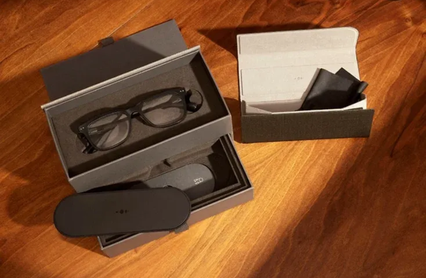 Las cajas abiertas revelan unas gafas inteligentes, un paño de limpieza y un estuche sobre una superficie de madera.