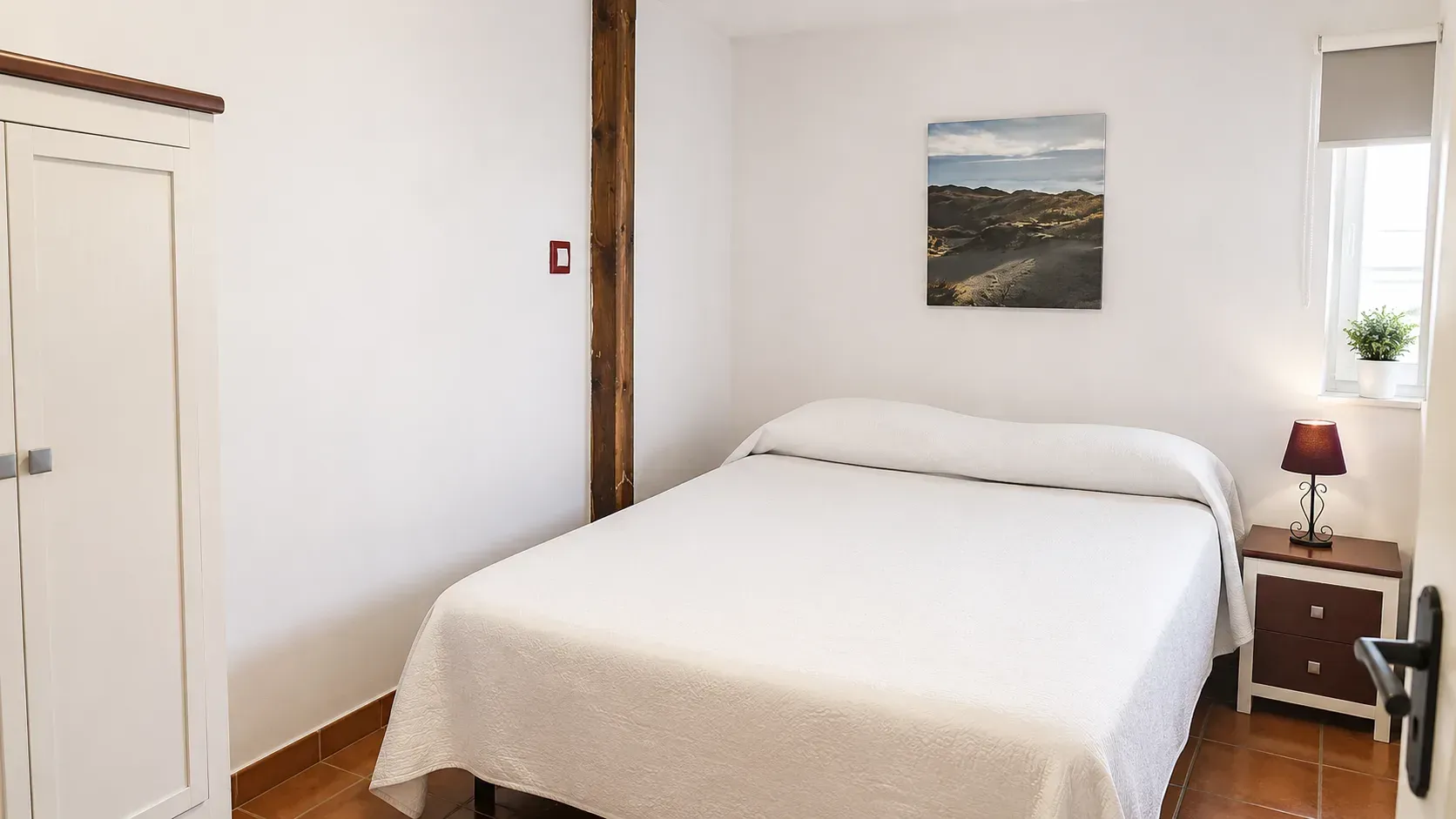 Habitación sencilla con cama blanca, lámpara de mesilla de noche, armario y cuadro mural con paisaje.