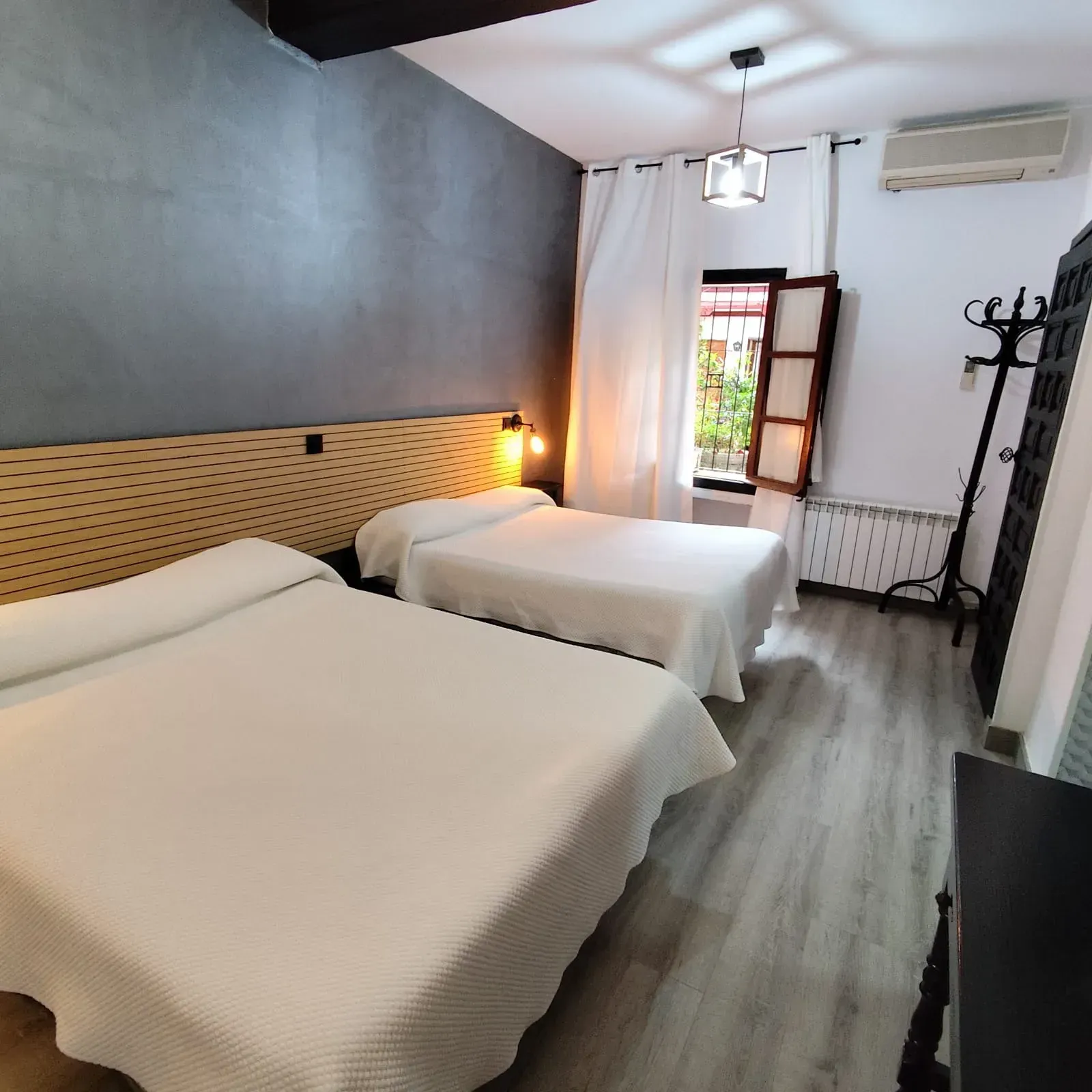 Acogedora habitación doble con dos camas blancas, lámpara cálida, ventana abierta y aire acondicionado de pared.