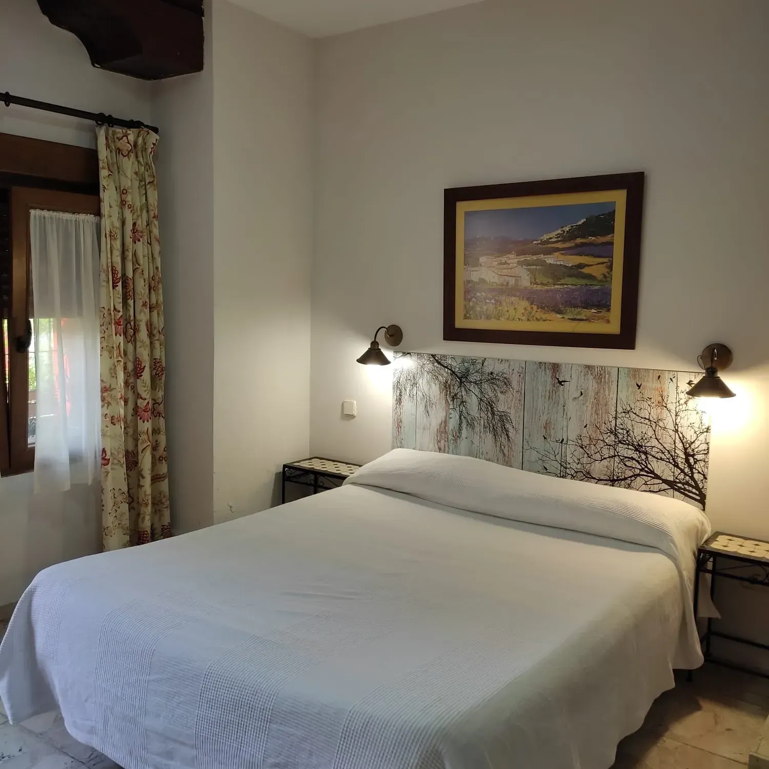 Habitación de hotel con una cama bien hecha, lámparas de pared y un paisaje enmarcado sobre el cabecero.