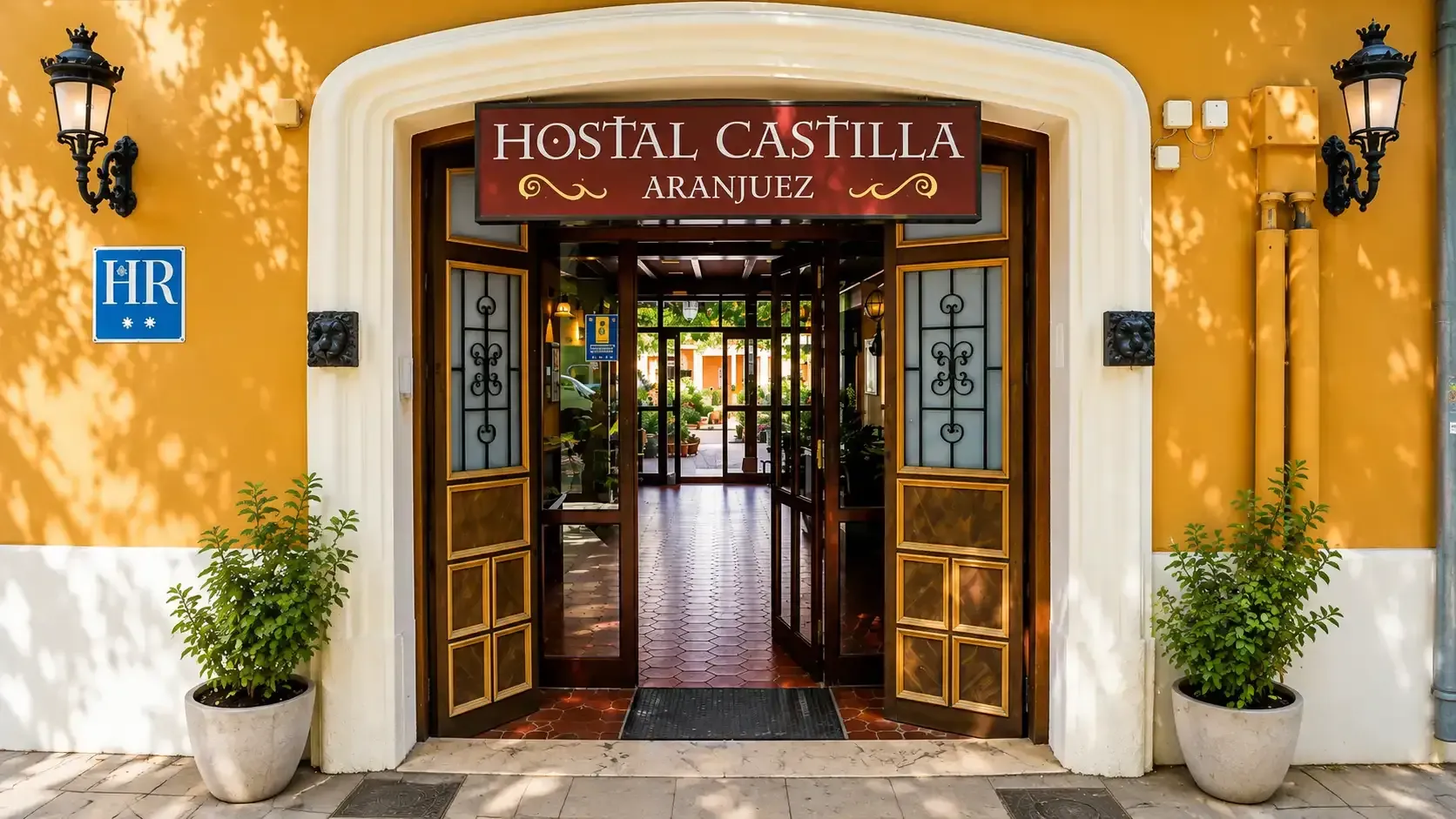 Entrada del Hostal Castilla, un edificio amarillo con puertas de madera abiertas y plantas en macetas junto a la entrada.