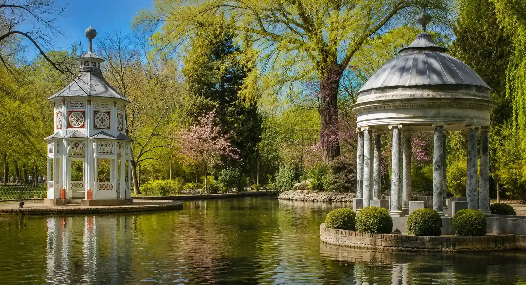 Dos cenadores blancos junto a un estanque tranquilo en un parque soleado con árboles primaverales.