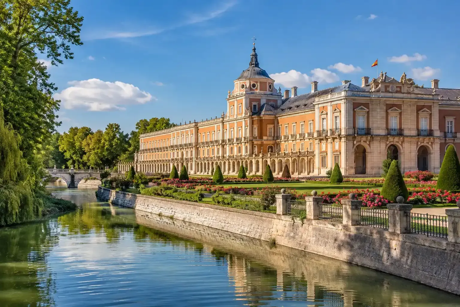 Palacio histórico junto a un río tranquilo, con una fachada de piedra ornamentada, cielo azul y árboles a lo largo del agua.