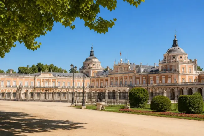 Gran fachada de palacio con cúpulas y arcos ornamentados, vista desde un patio arenoso bajo árboles frondosos.