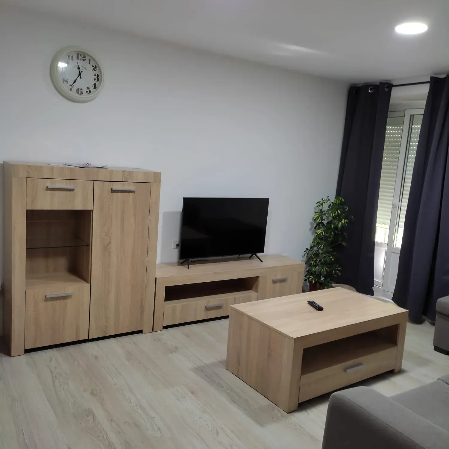 Sala de estar moderna con muebles de madera clara, televisor, mesa de centro, reloj de pared, planta y cortinas oscuras.