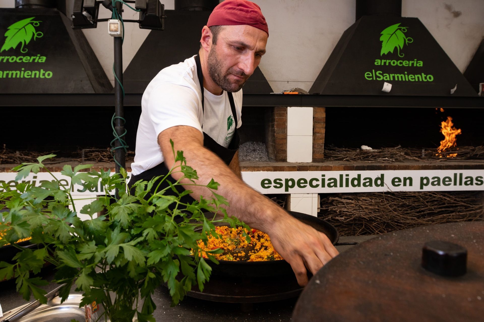 Un hombre está cocinando comida en una sartén grande frente a un cartel que dice especialidad en paellas.