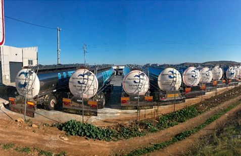 Una hilera de camiones cisterna plateados estacionados en un terreno de tierra bajo un cielo azul.