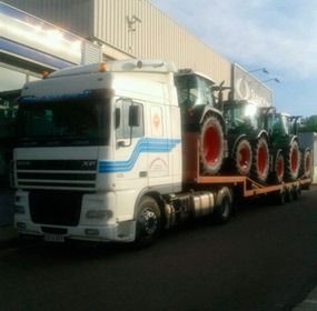 Un camión con remolque blanco transporta tractores rojos en un camión de plataforma en una carretera cerca de edificios.