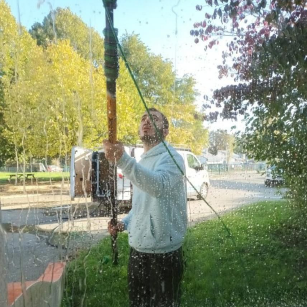 Une personne utilise une brosse à long manche pour laver une fenêtre à l'extérieur, avec des arbres verts en arrière-plan.