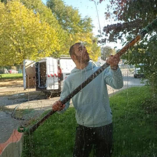 Un homme lave une vitre avec un outil à long manche, debout à l'extérieur près d'un camion stationné et d'arbres.