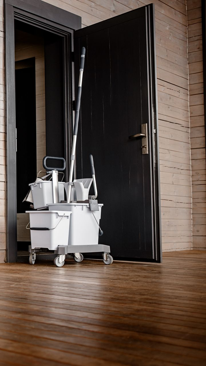 Chariot de nettoyage avec balai et seaux près d'une porte noire ouverte sur un sol en bois.