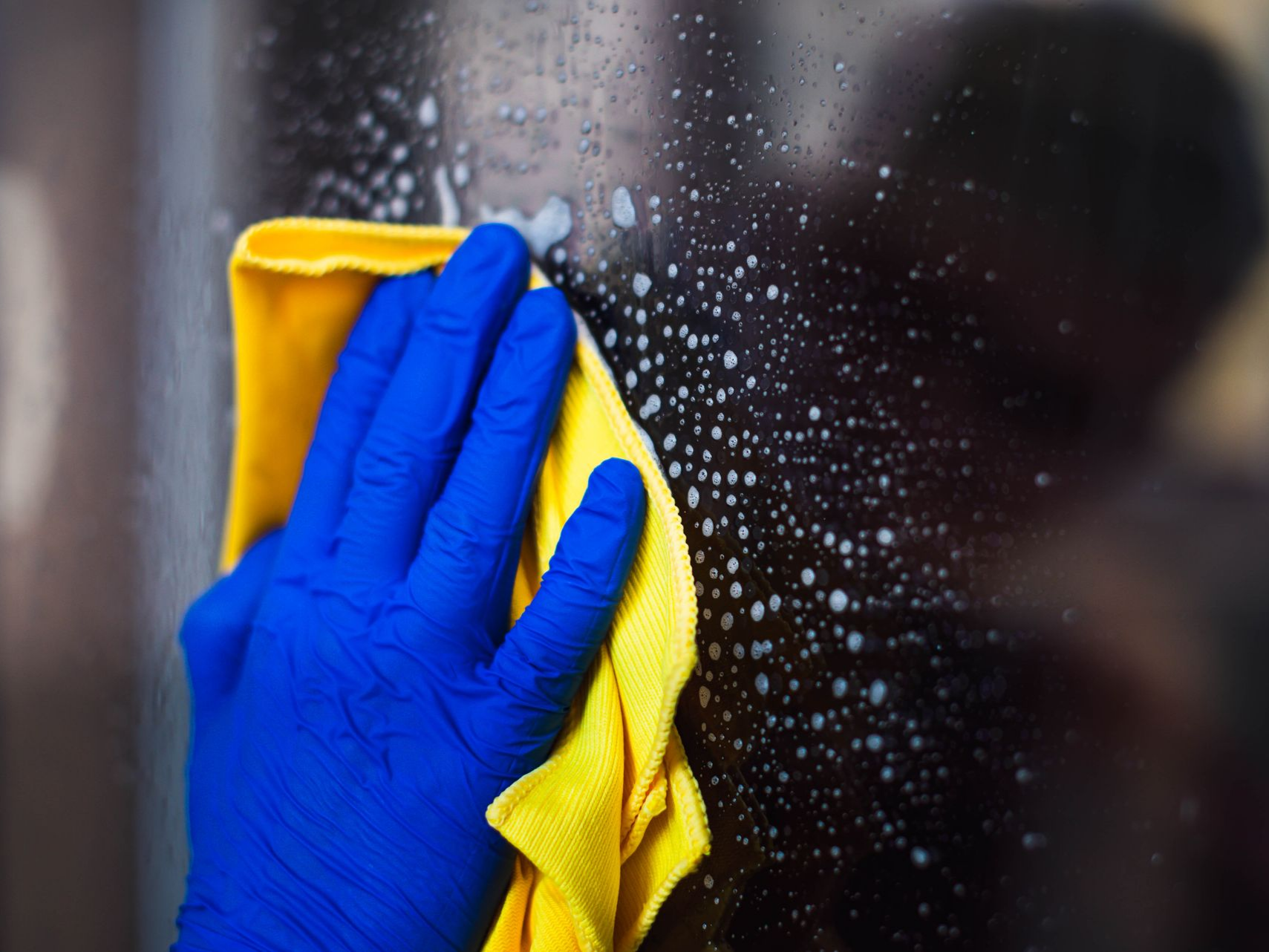 Une main gantée bleue essuie une vitre savonneuse avec un chiffon jaune.