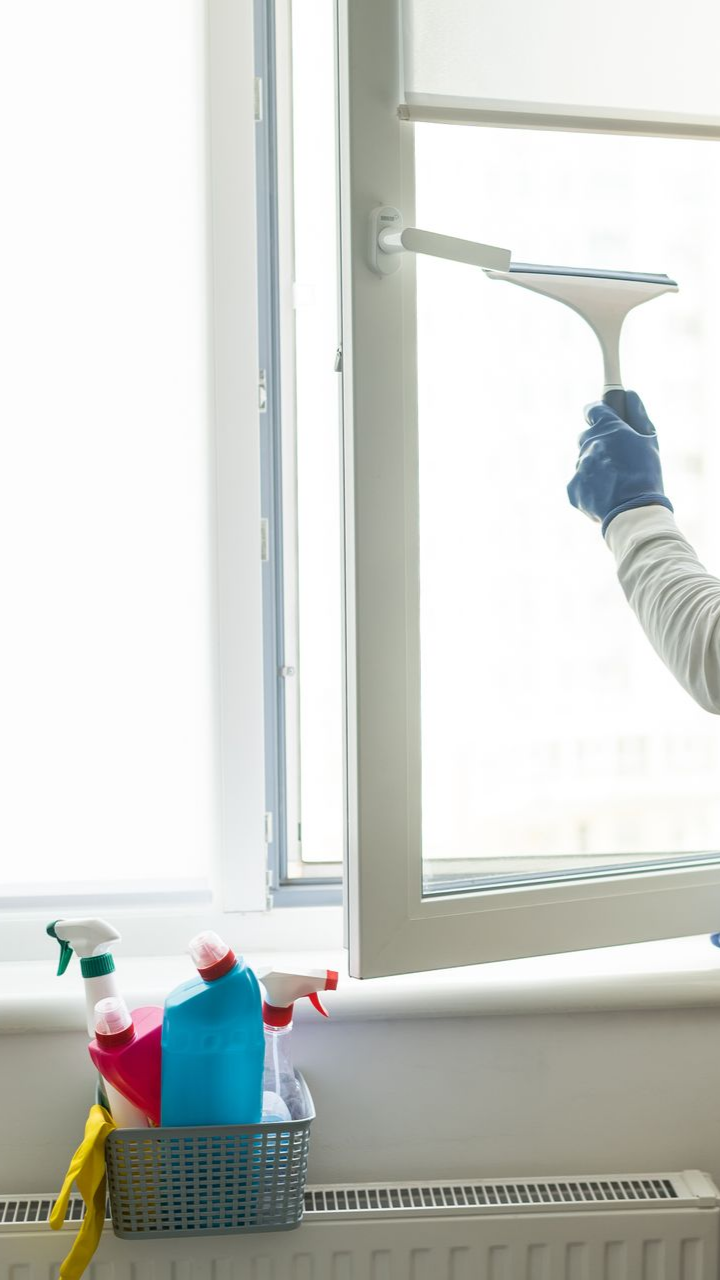 Une personne nettoie une vitre avec une raclette ; des produits de nettoyage sont rangés dans un panier sur le rebord de la fenêtre.