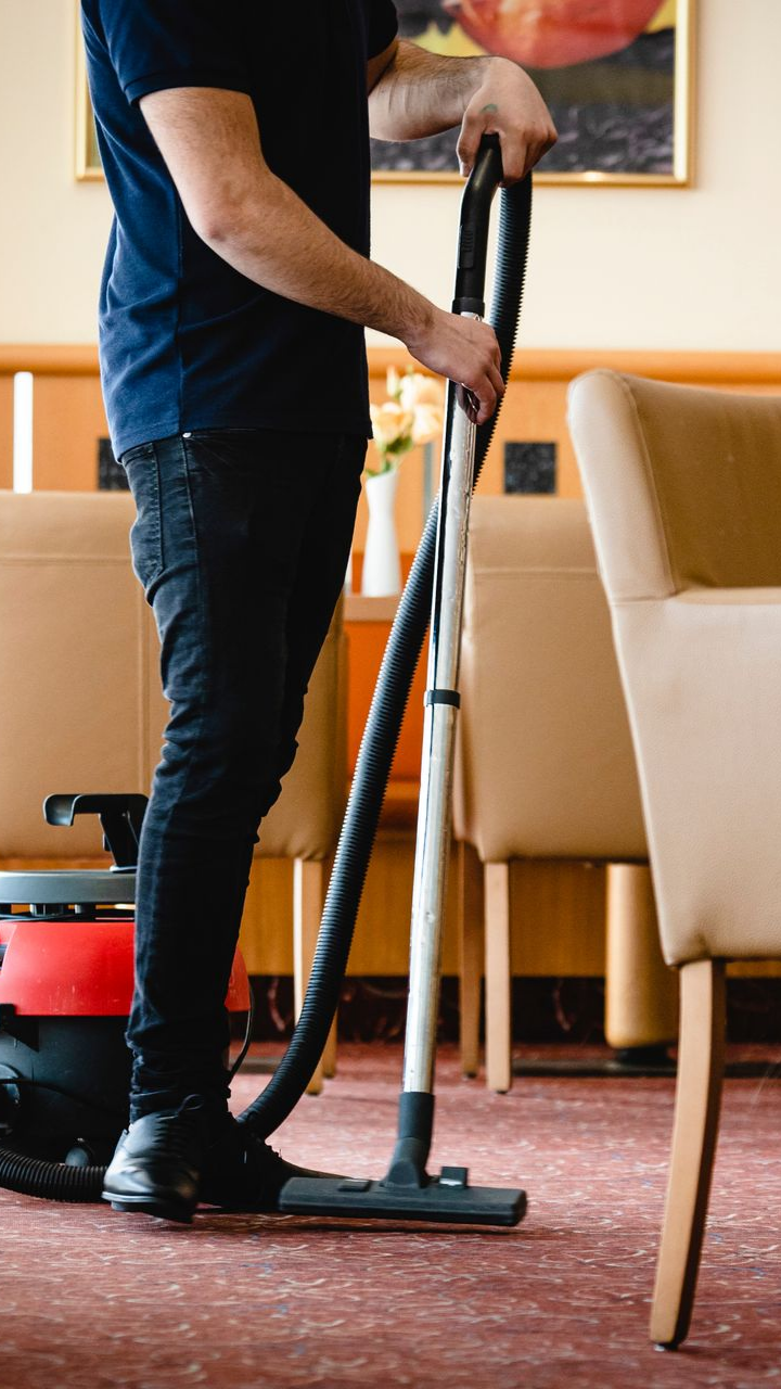 Une personne passe l'aspirateur sur une moquette rouge dans une pièce meublée de chaises beiges et utilisant un aspirateur rouge et noir.