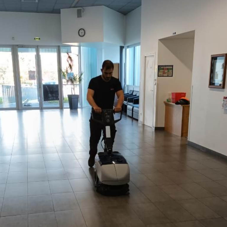 Une personne utilise une autolaveuse dans le couloir d'un immeuble, nettoyant un sol carrelé.