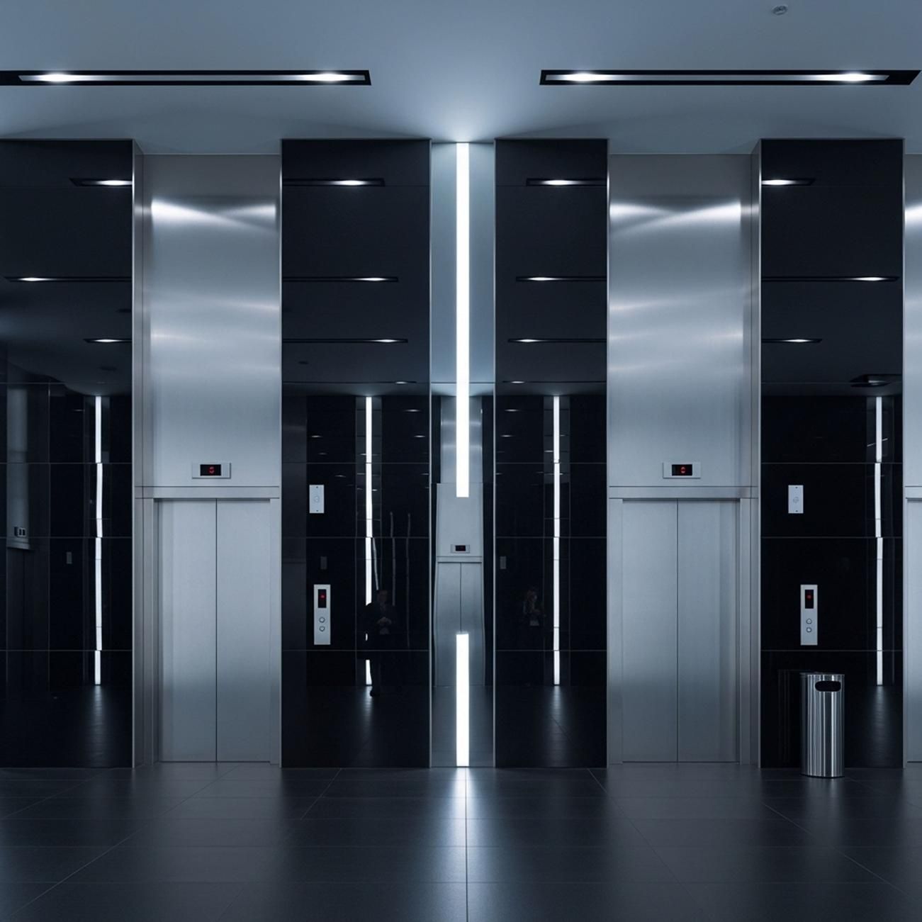 Vestíbulo de ascensor moderno con puertas negras y plateadas brillantes bajo luces cenitales brillantes.