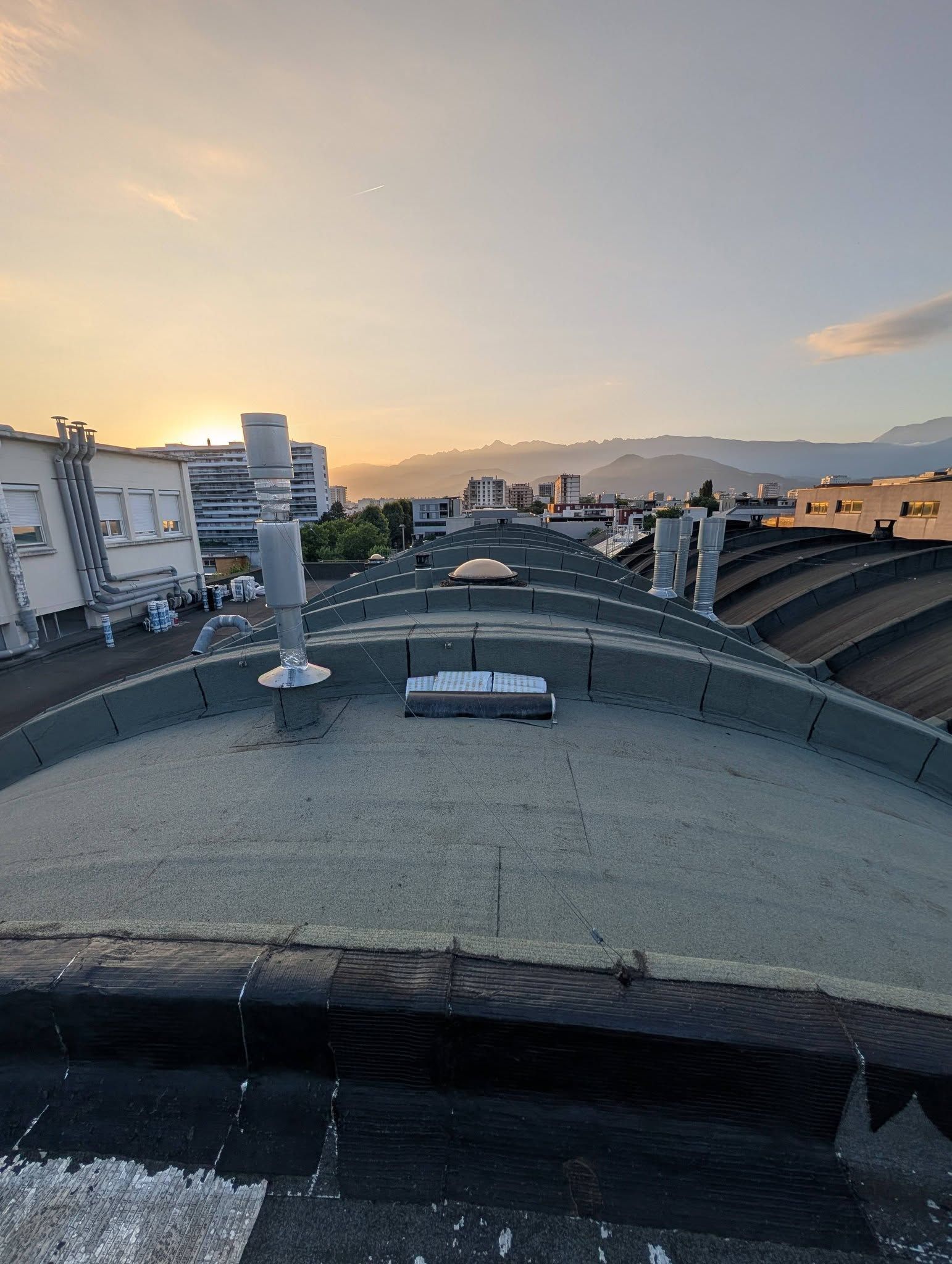 Vue depuis un toit incurvé recouvert de gravier au coucher du soleil, avec des montagnes et des bâtiments urbains au loin.