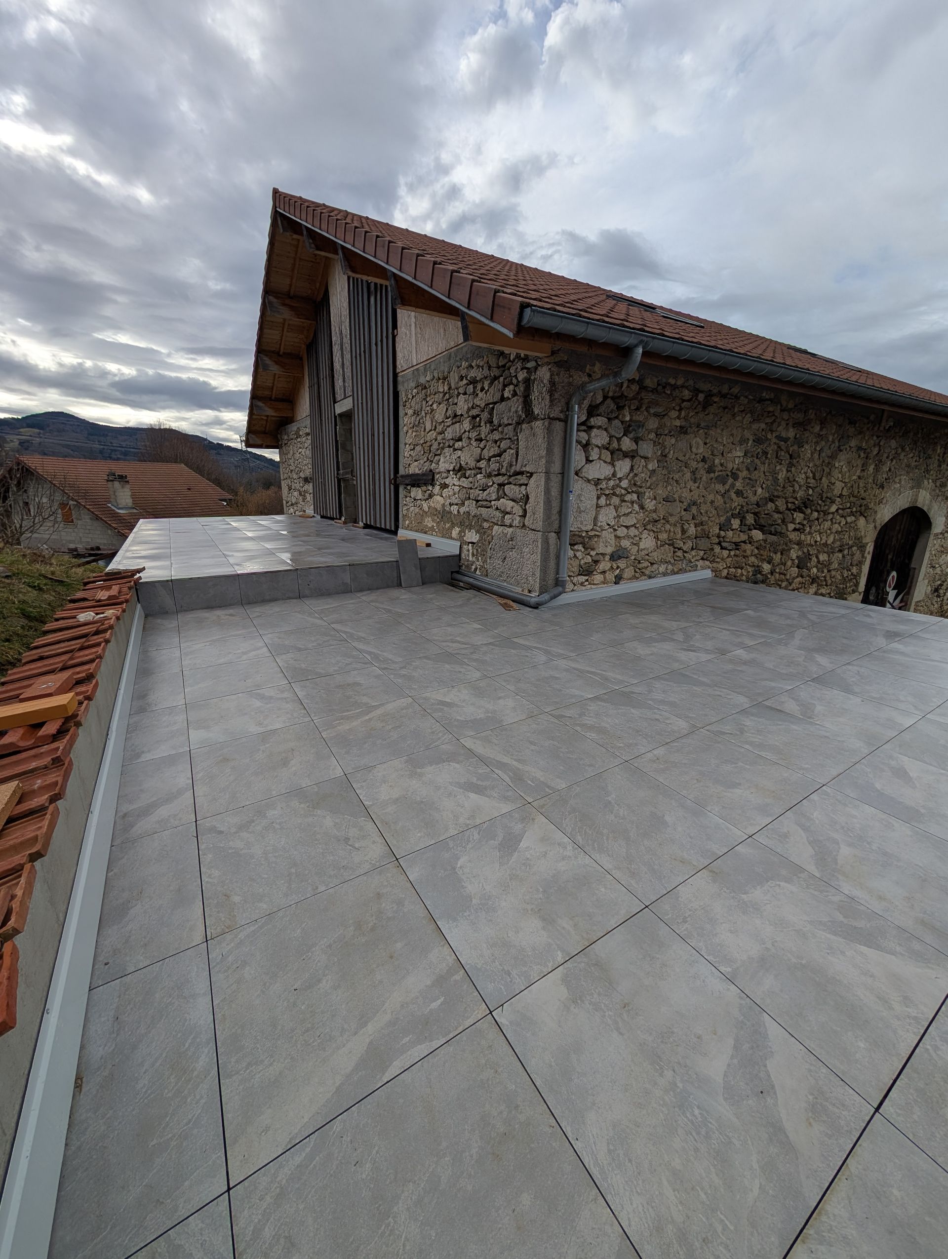 Une terrasse en carrelage gris fraîchement posée devant une grange rustique en pierre.