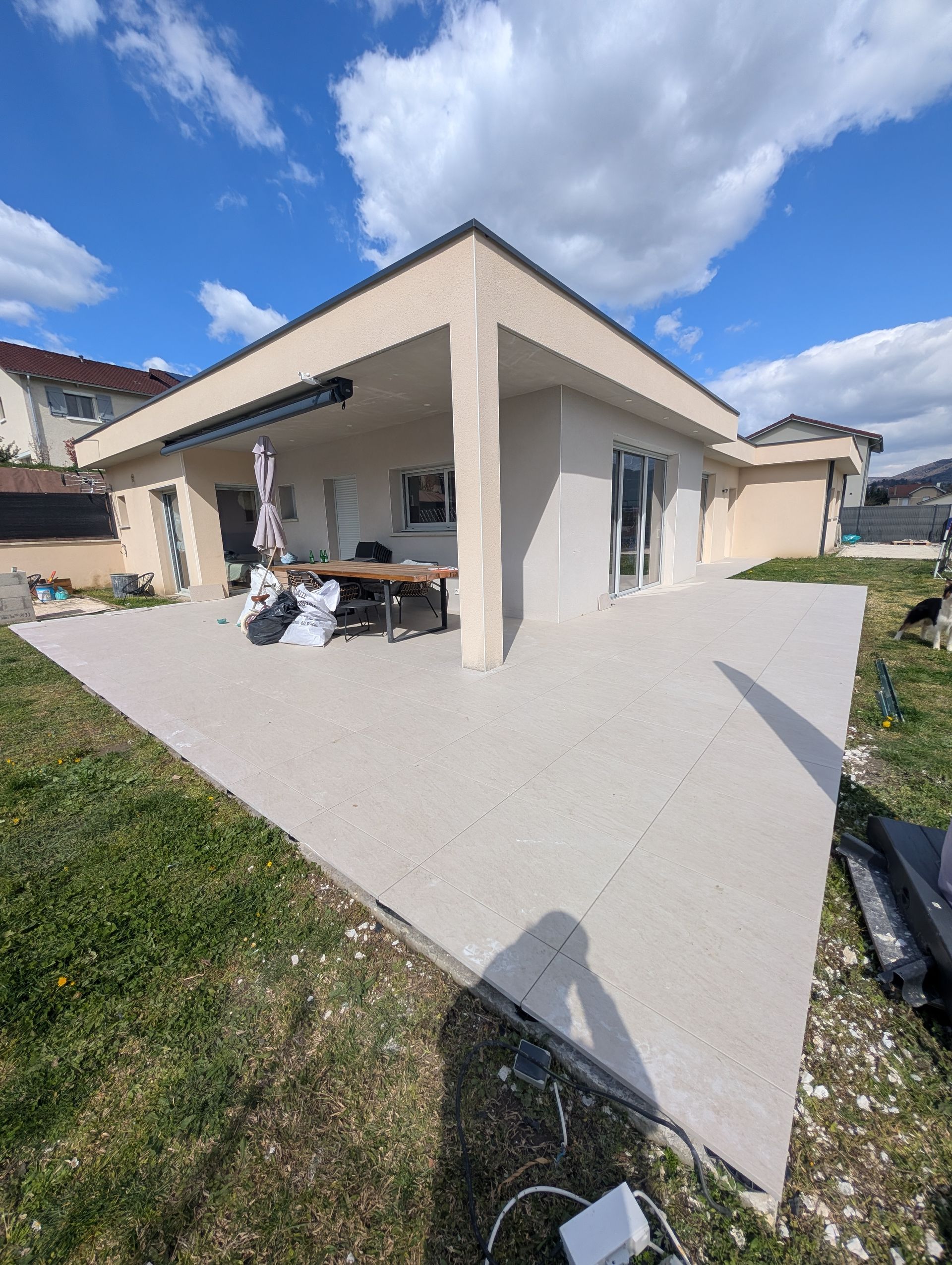 Une maison moderne aux tons clairs avec une grande terrasse, un espace repas extérieur et une pelouse.