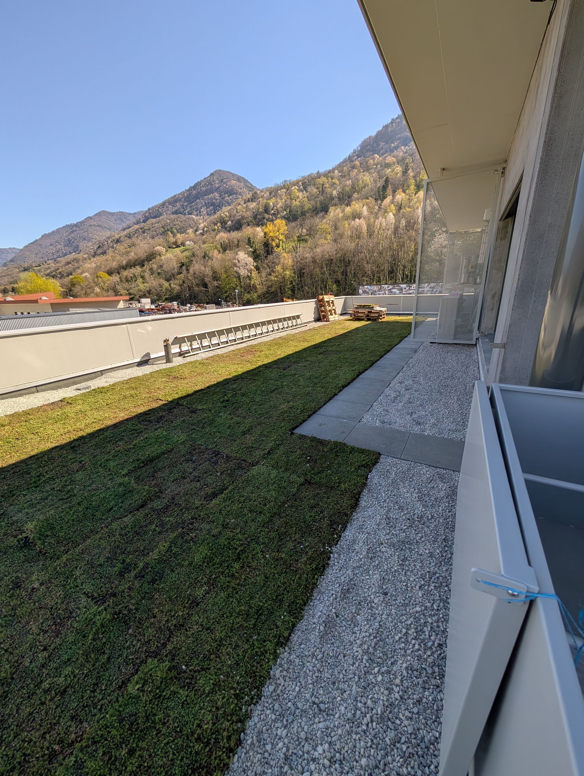 Vue sur une terrasse en hauteur avec une pelouse, un chemin de gravier blanc et un paysage montagneux en arrière-plan.