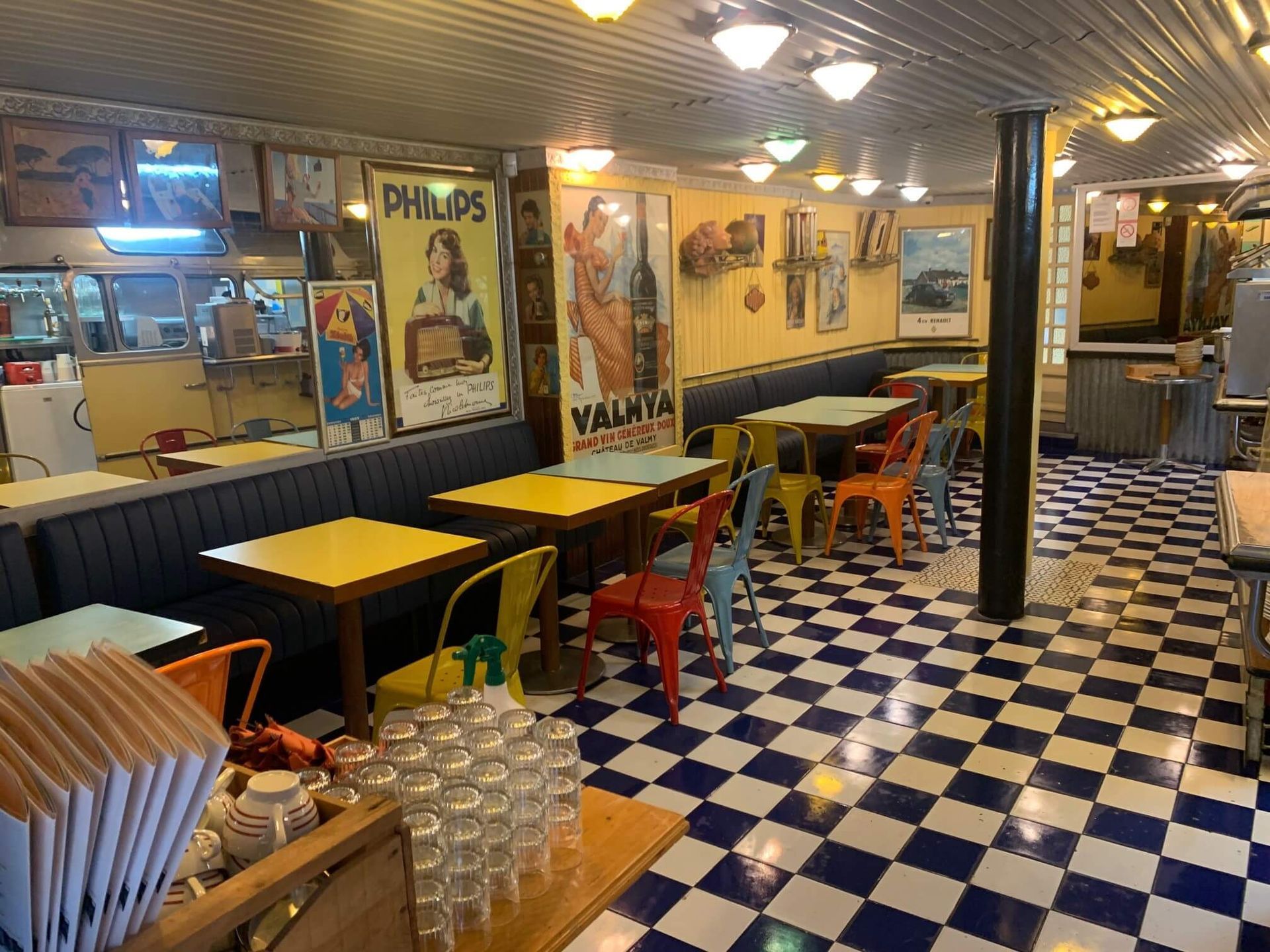 Intérieur d'un restaurant rétro : sol à damier, tables et chaises colorées, affiches vintage aux murs.
