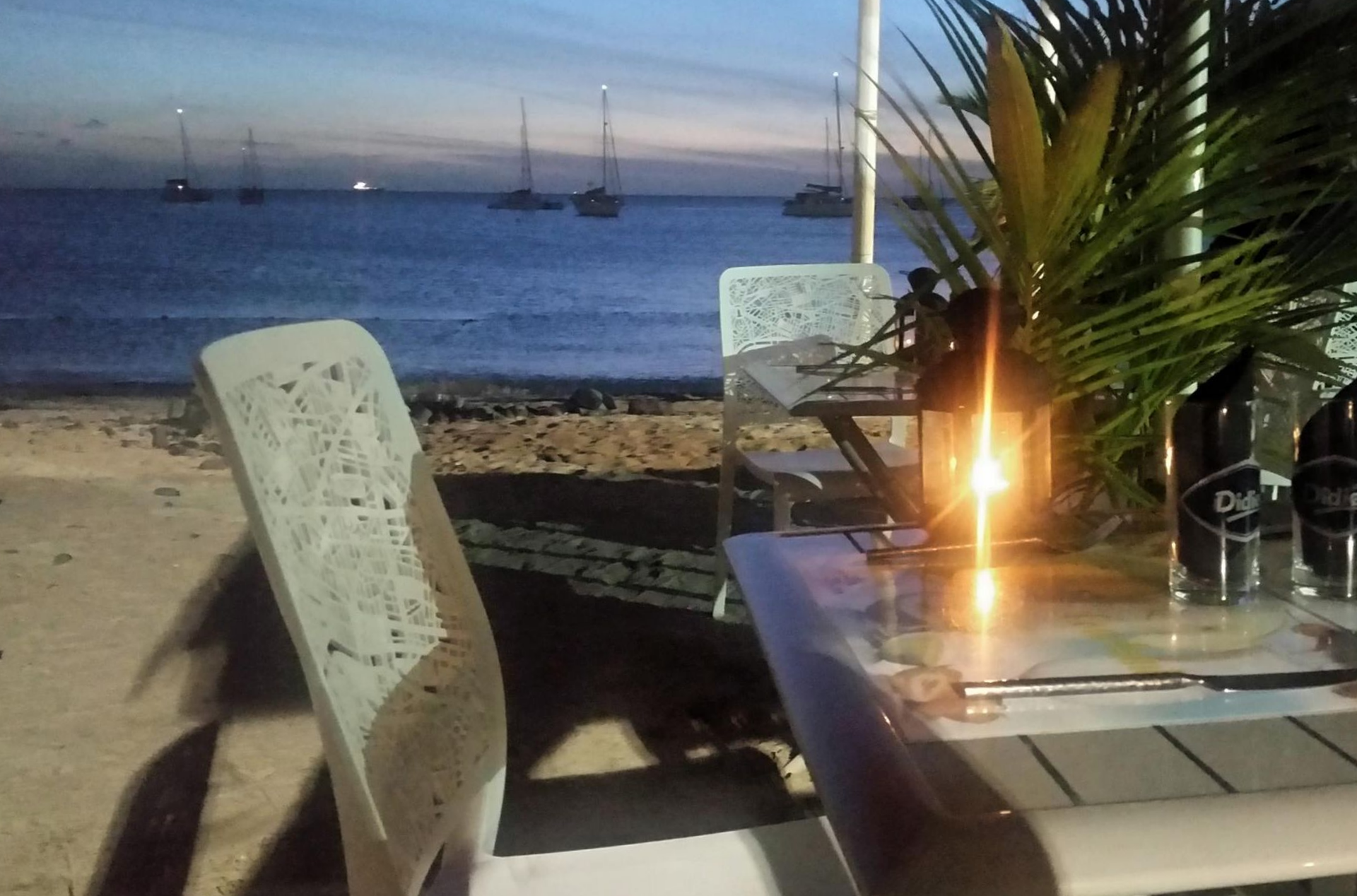 Table dressée pour le dîner dans un restaurant en bord de mer, éclairée à la bougie. L'océan et les voiliers se dessinent au crépuscule.