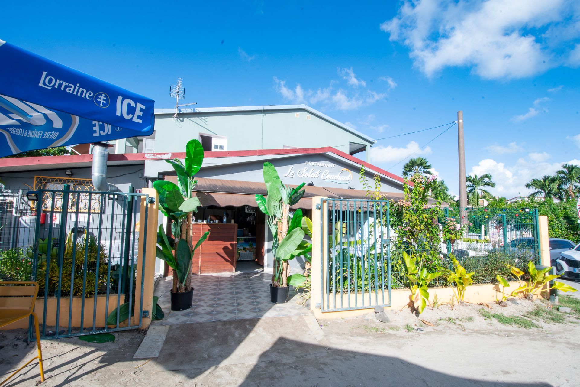 Un petit bâtiment avec une entrée carrelée, un portail en fer forgé et des plantes d'extérieur. On aperçoit un parasol ICE.