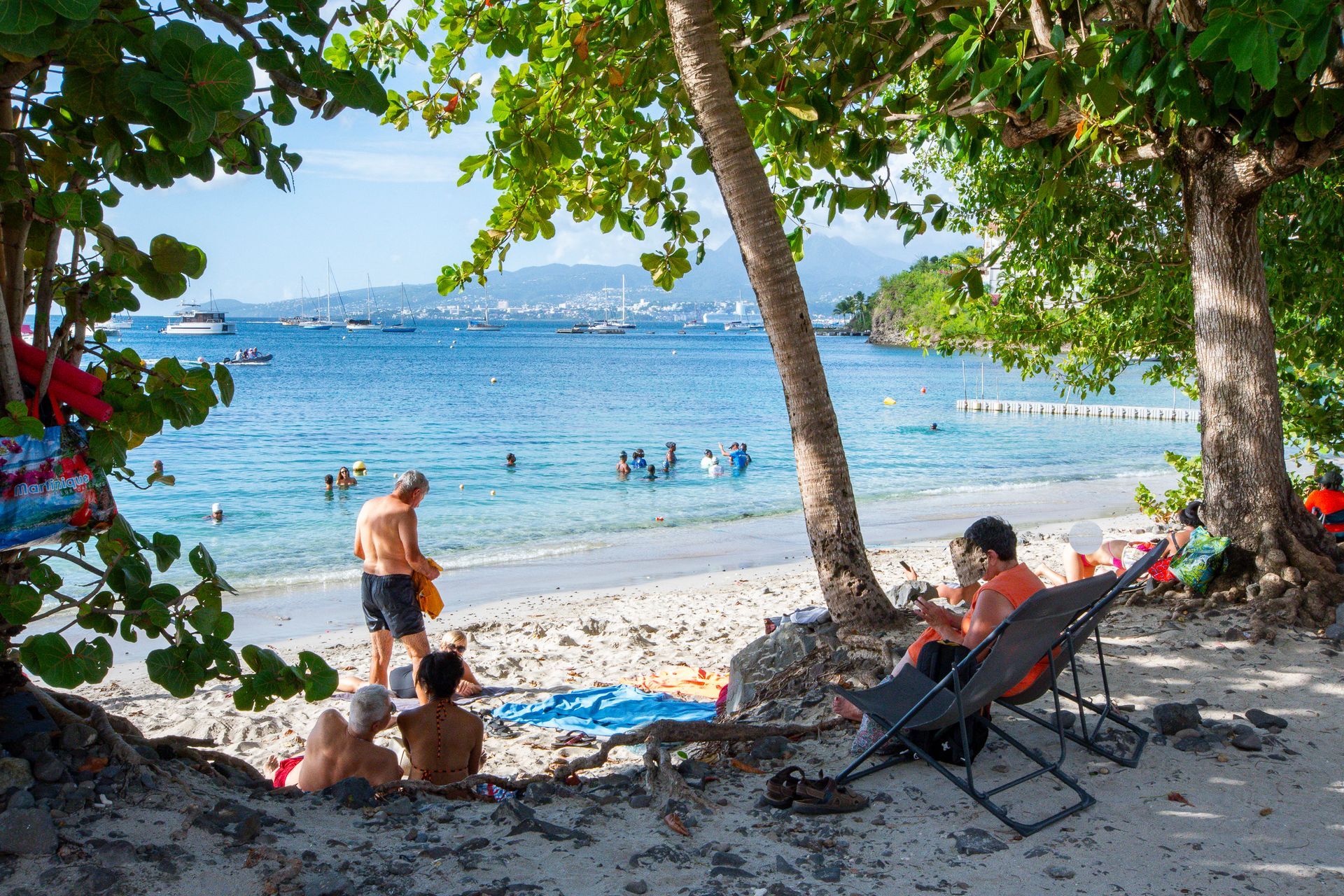 Scène de plage : des gens se baignent dans l’eau bleue ; d’autres se détendent à l’ombre. Des bateaux naviguent sur la mer, une végétation luxuriante borde le rivage.