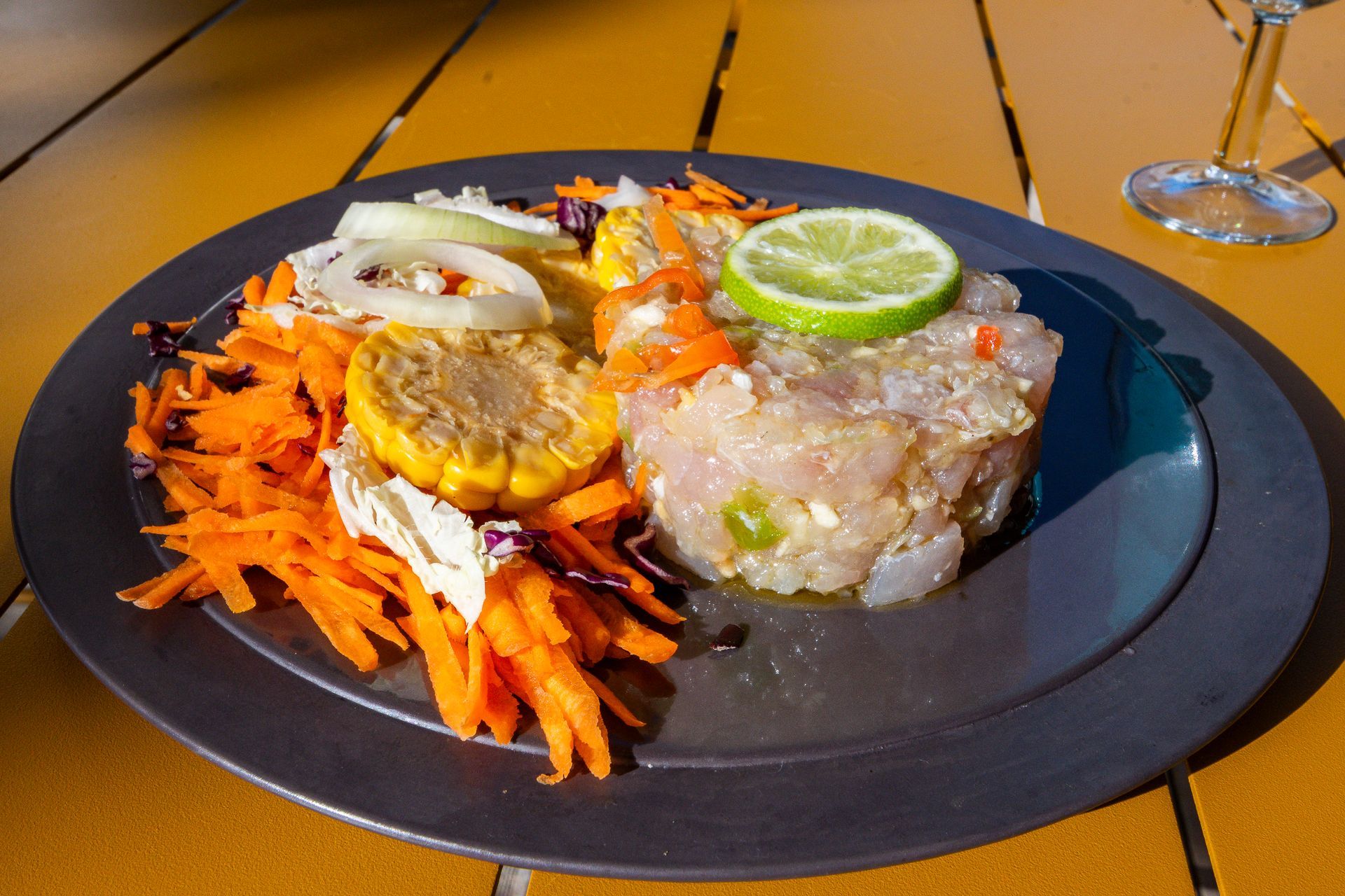 Tartare de poisson dans une assiette foncée avec des carottes râpées, du maïs, des oignons et une tranche de citron vert, sur une table en terrasse.