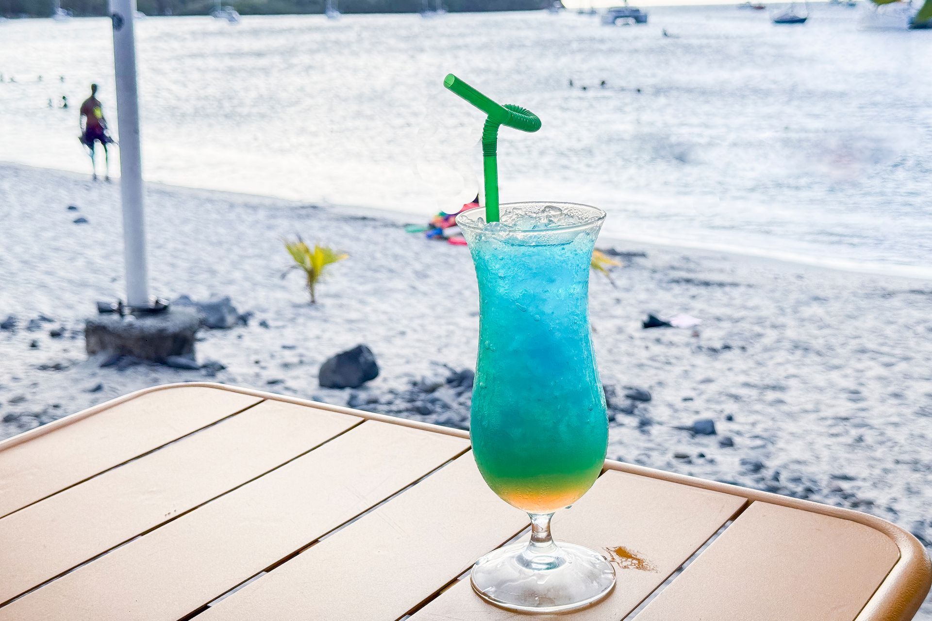 Cocktail bleu sur une table donnant sur une plage ; paille verte, verre transparent, plage de sable en arrière-plan.