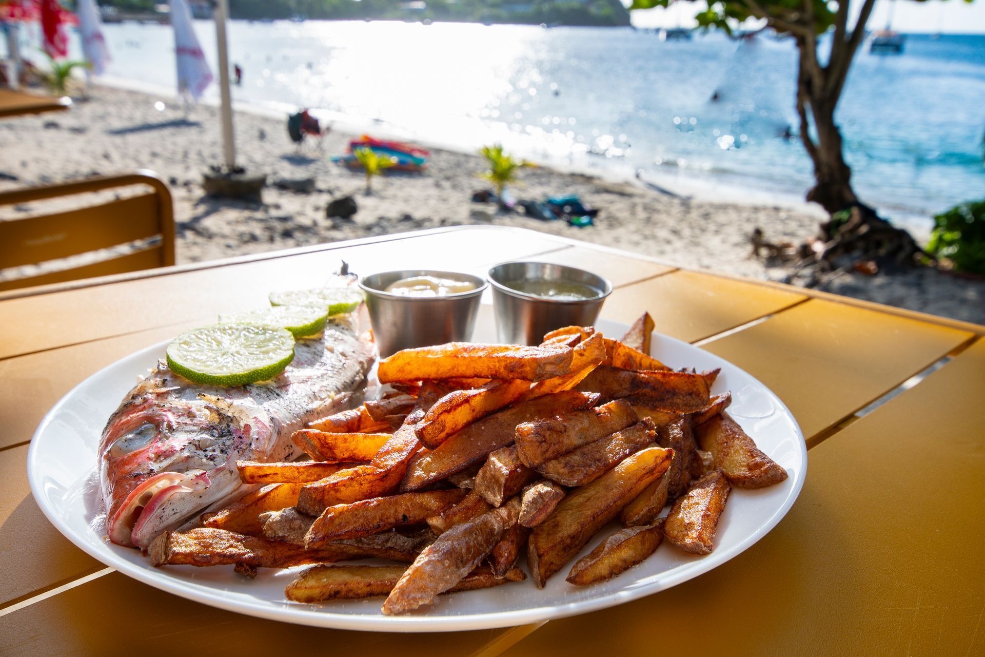 Poisson grillé avec frites, citron vert et sauces, servi sur une assiette blanche, sur fond de plage.