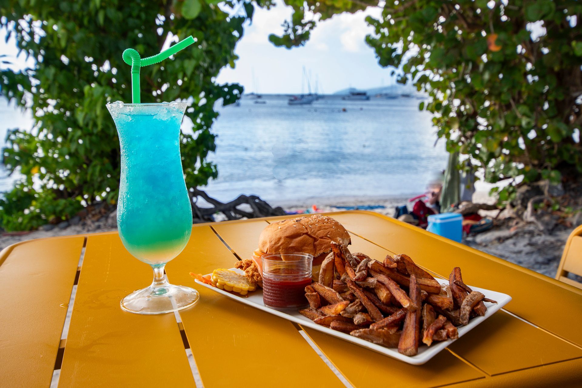 Cocktail bleu, hamburger et frites sur une table jaune en bord de plage.